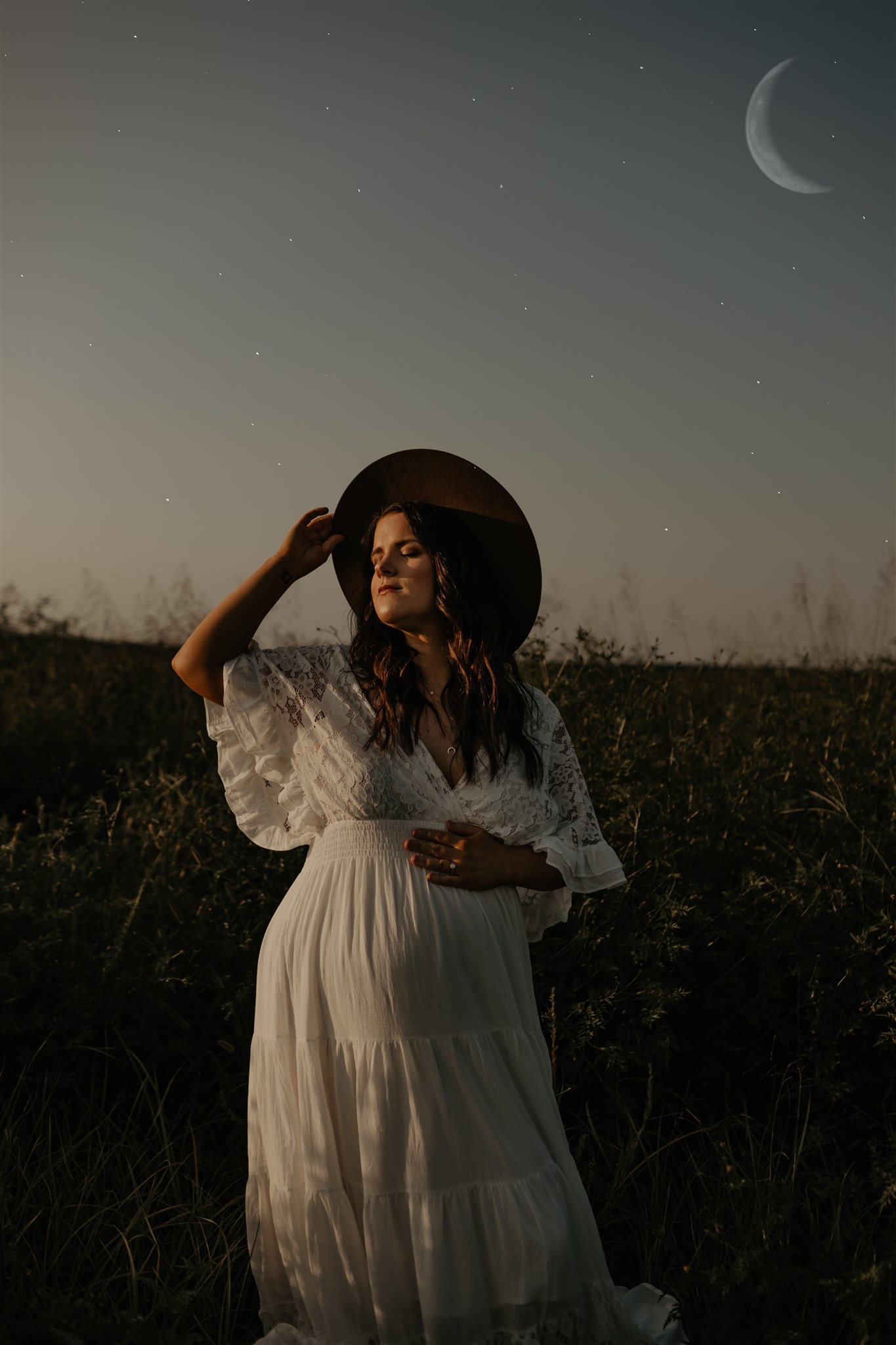 Serene pregnant woman in sunset field with the moon in the background. She has a hand on her belly and on her hat brim