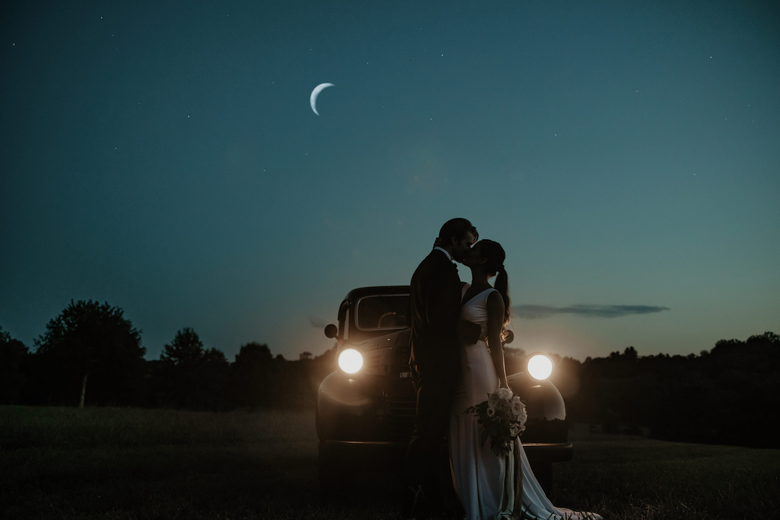 Dusk picture of bride and groom kissing in front of the hood of the vintage pick up. Its headlights are on and a crescent moon in the sky